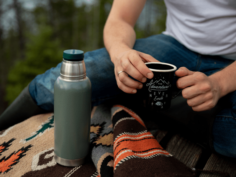 a woman holding a cup of coffee while sitting on a log while camping