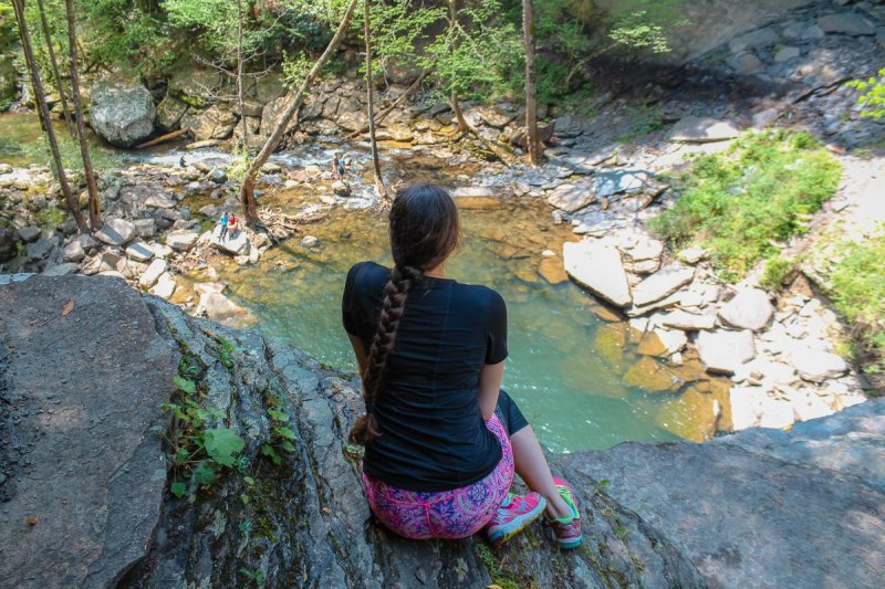 a woman sitting on the edge of a waterfall cliff looking down below during a hike in Tennessee.